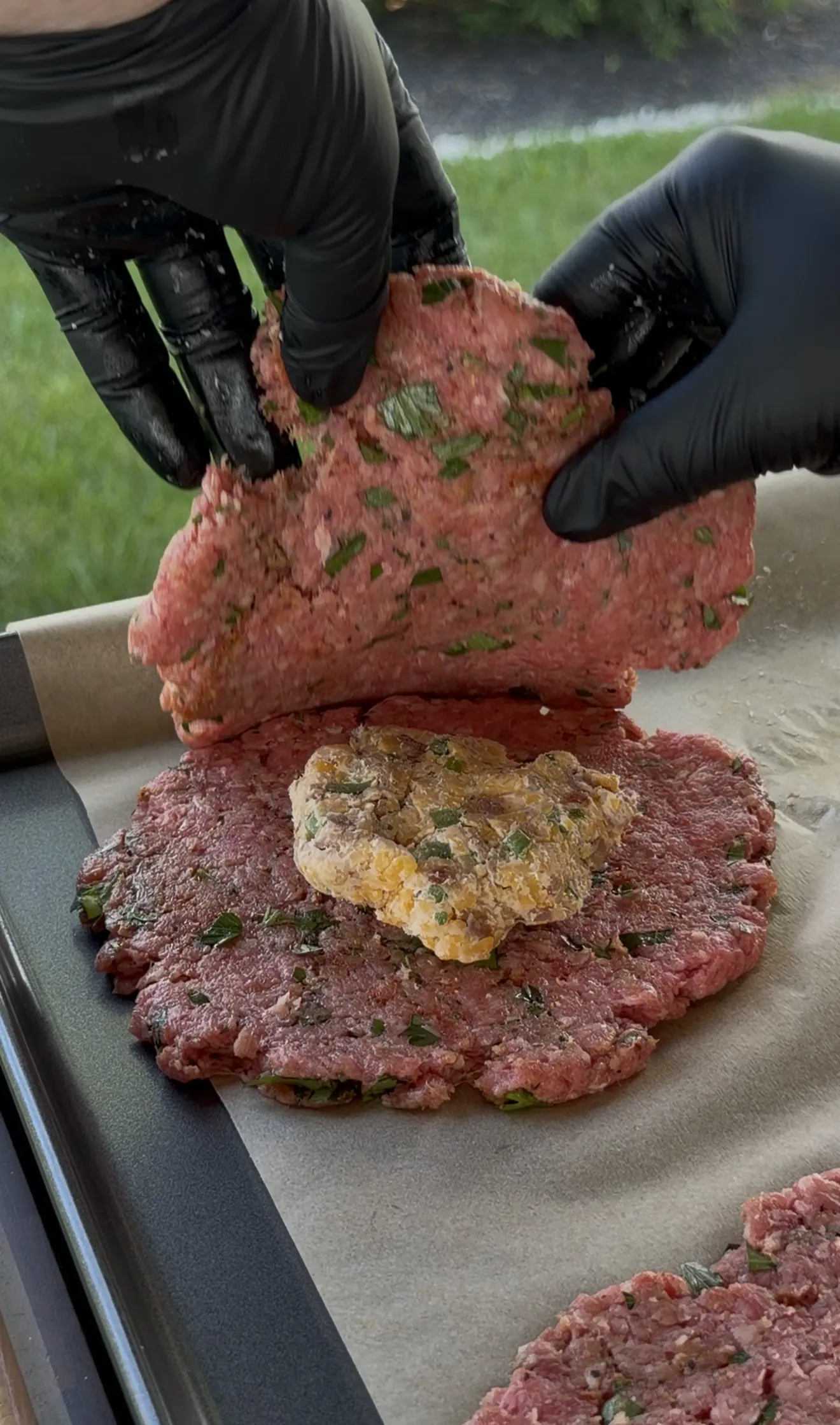 Gloved hands sealing a stuffed burger patty around the cheese filling on parchment
