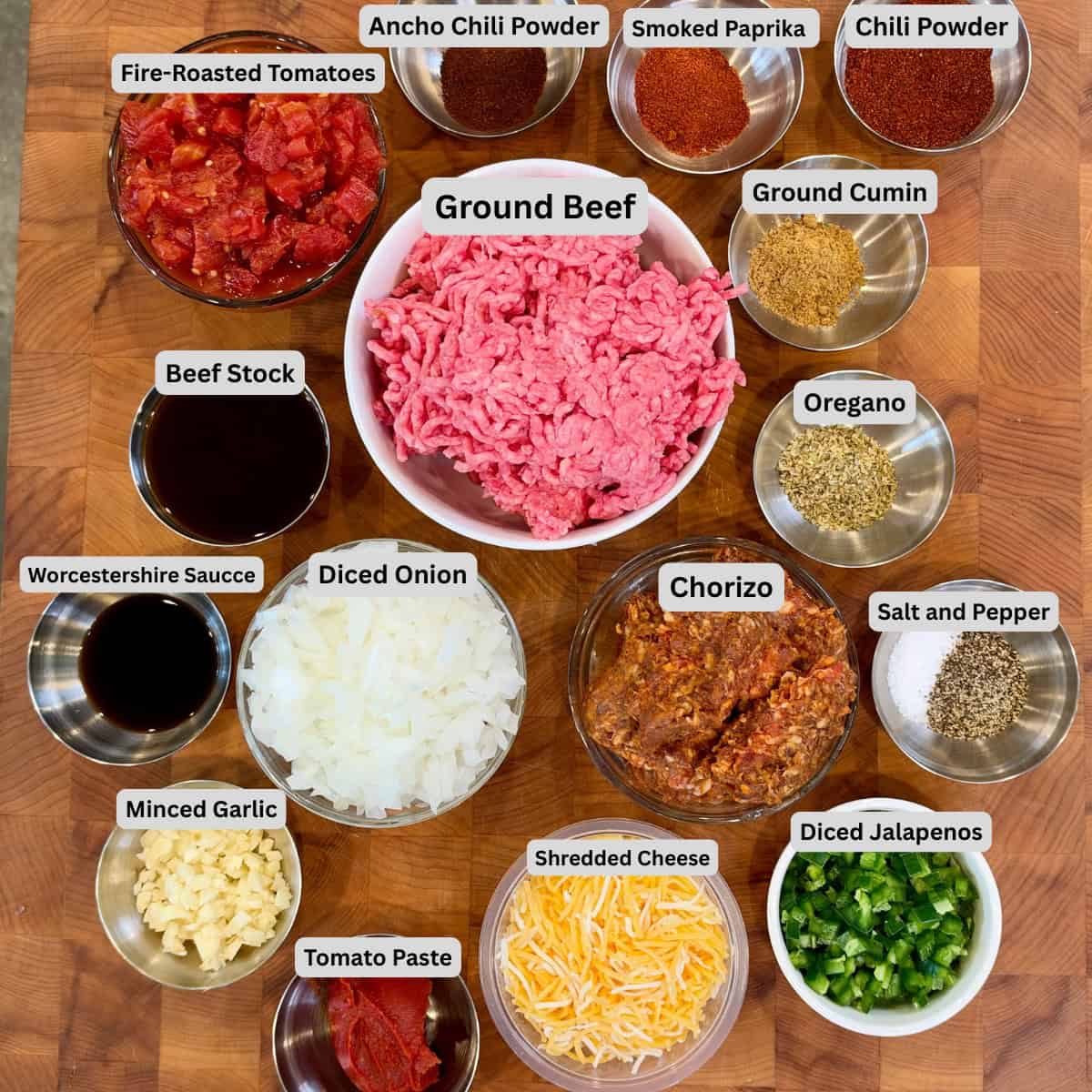 Labeled ingredients for an easy chili recipe with ground beef, Mexican chorizo, fire-roasted tomatoes, beef stock, diced onion, jalapeños, garlic, tomato paste, Worcestershire sauce, and chili spices.