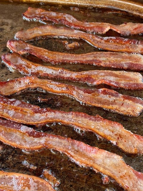 Crispy bacon strips sizzling in rendered bacon fat on a flat-top griddle for Easy Breakfast Sliders.