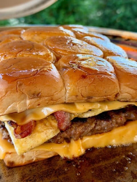 Easy Breakfast Sliders assembled on a flat-top griddle with a large sausage patty, folded eggs, crispy bacon, melted American cheese, and maple-Dijon glazed Hawaiian rolls.