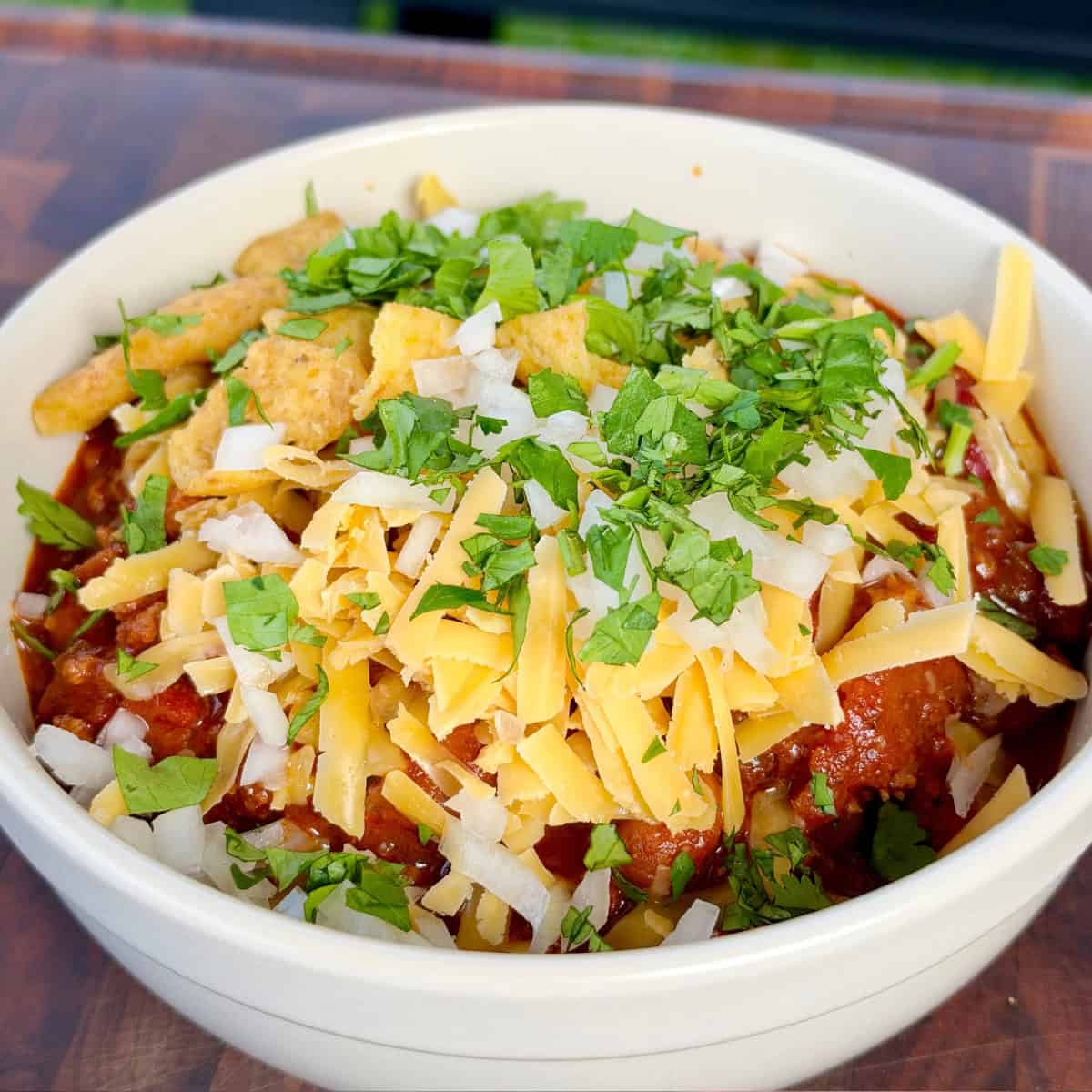 Smoked Over the Top Chili topped with shredded cheddar, diced onion, cilantro, and corn chips
