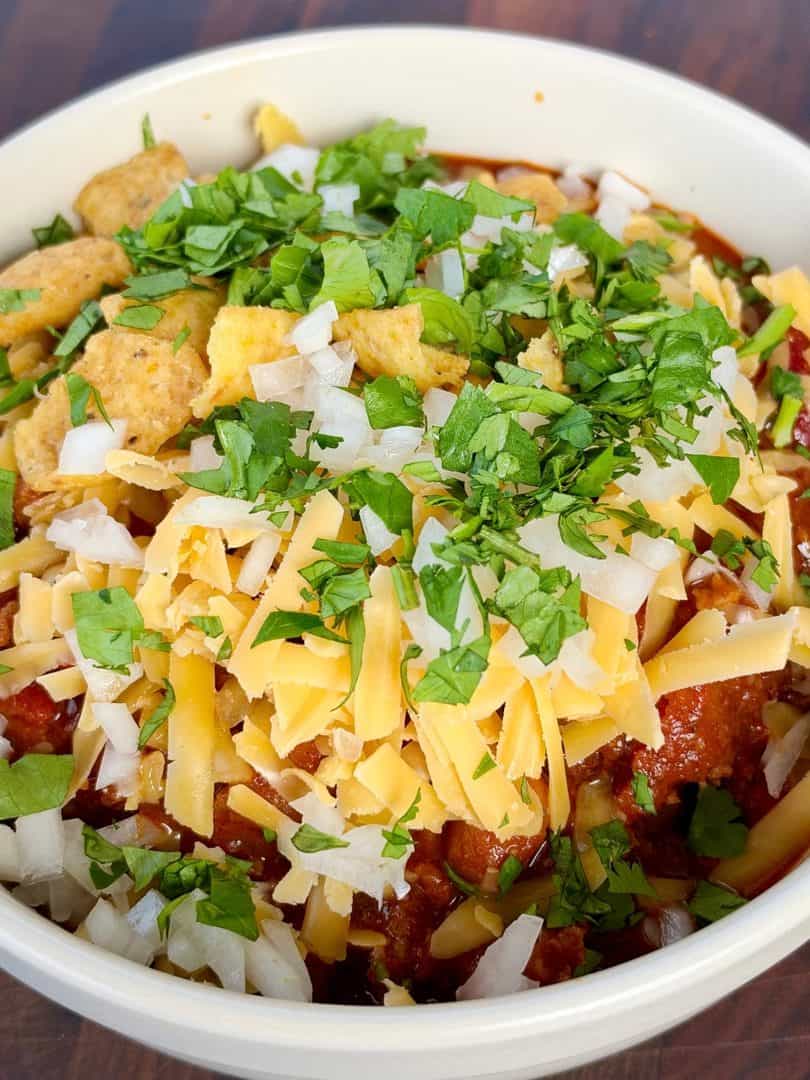 Smoked Over the Top Chili topped with shredded cheddar, diced white onion, cilantro, and corn chips.