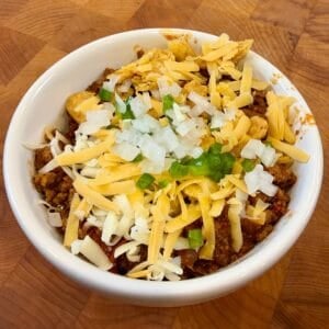 Easy chili recipe with ground beef and Mexican chorizo topped with shredded cheese, diced onions, jalapeños, and crunchy corn chips in a white bowl.