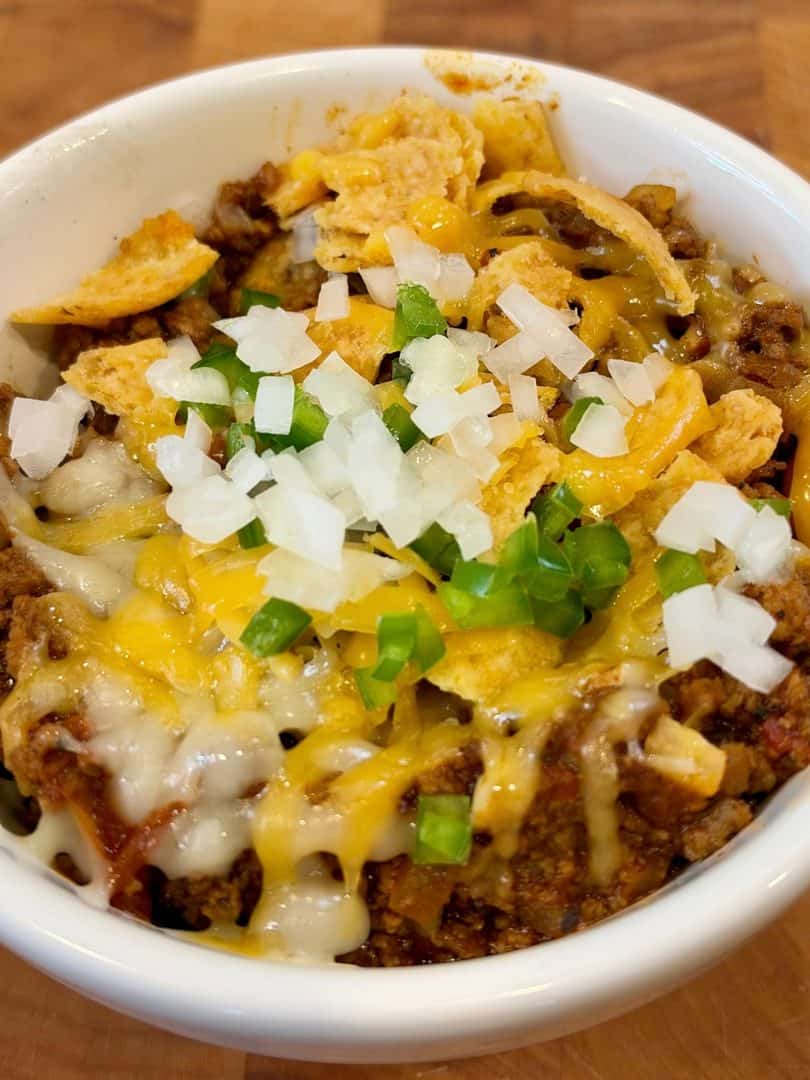 Easy chili recipe with ground beef and Mexican chorizo topped with shredded cheese, diced onions, jalapeños, and crunchy corn chips in a white bowl.