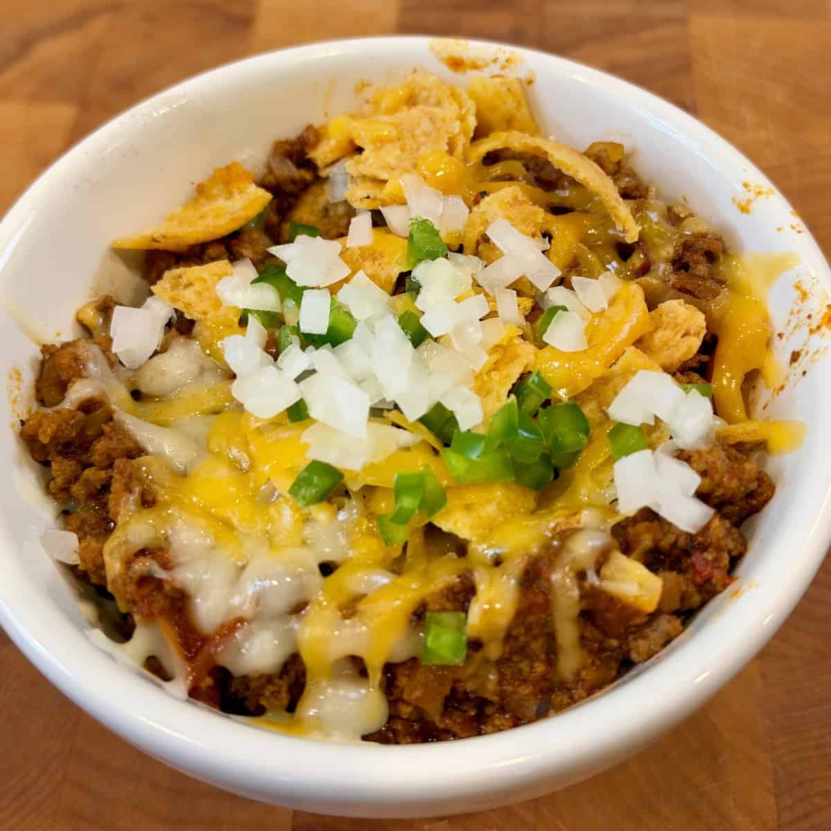 Easy chili recipe with ground beef and chorizo topped with melted shredded cheese, diced white onions, jalapeños, and crunch corn chips in a while bowl. 