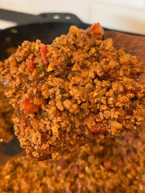 Close up of thick, no-bean chili made with ground chuck, Mexican chorizo, fire-roasted tomatoes, onions, and Jalapeños on a wooden spoon.