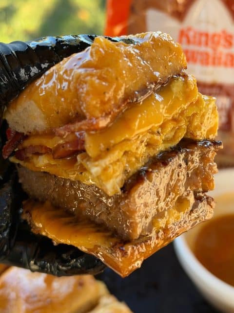 Easy Breakfast Slider held in a gloved hand, showing crispy bacon, folded eggs, a sausage patty, and melted American cheese on a maple-Dijon glazed Hawaiian roll.